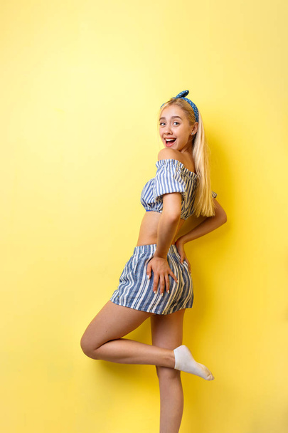 Joyful beautiful blonde girl in summer outfit is smiling standing in a half-turn, lifting one leg up, over yellow background. Emotions, gestures and facial expression - 写真・画像