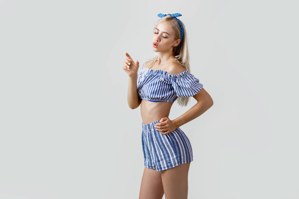 Beautiful blonde girl in summer outfit, posing at camera holding something like a bottle in her hand and drinks it, standing over white background - Valokuva, kuva
