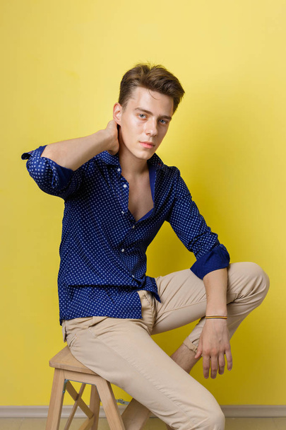 Calm handsome dark-haired guy wearing blue shirt and beige pants sitting on wooden stool over yellow wall - Φωτογραφία, εικόνα