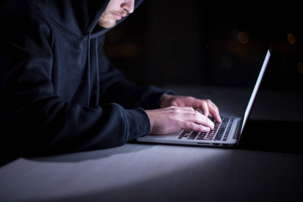 Young talented hacker using laptop computer while working in dark office with big city lights in the background at night - Photo, Image