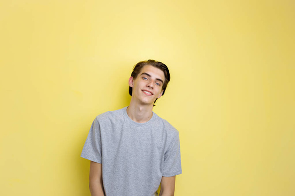 Cheerful handsome young thin dark-haired guy with blue eyes wearing gray t-shirt, standing against yellow background - Foto, afbeelding