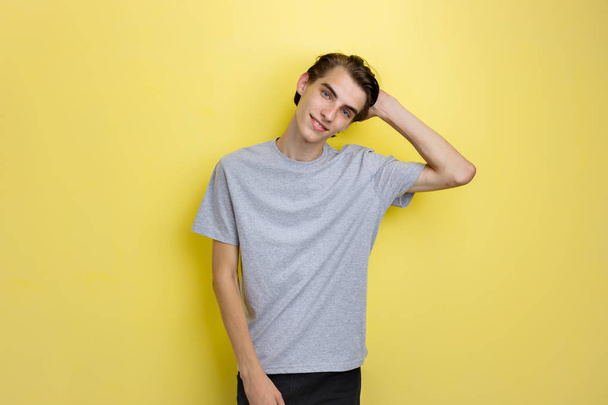 Cheerful handsome young thin dark-haired guy with blue eyes wearing gray t-shirt holding hand on head, standing against yellow background - Foto, imagen