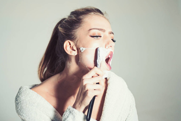 Woman with face covered with foam holds straight razor in hand. Girl on busy face wears bathrobe, grey background. Barber and shaving concept. Lady shaves her face with sharp blade of straight razor - Foto, immagini
