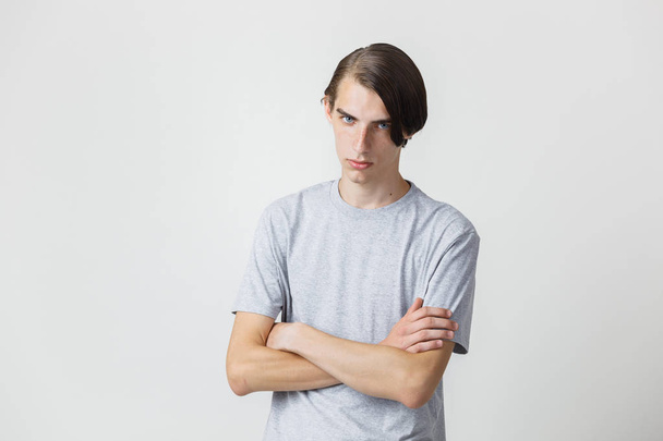 Very serious handsome young thin dark-haired guy with blue eyes wearing gray t-shirt standing against white background with crossed hands - Photo, Image
