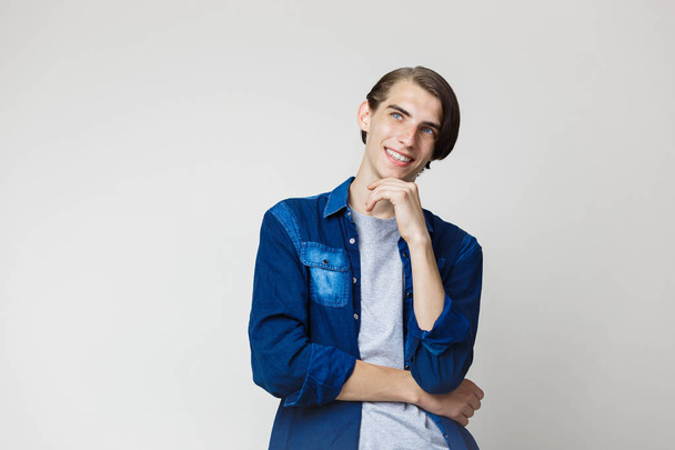 Smiling handsome young thin dark-haired guy with blue eyes wearing blue denim shirt, looking left and up, standing against white background - 写真・画像