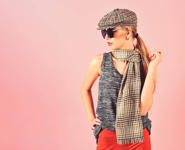 Side view skinny blond model in tartan beige cap and scarf and black sunglasses isolated on pink background. Girl in bright orange pants playing with her ponytail - Foto, Imagem