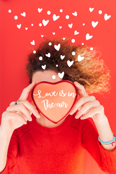 top view of girl holding heart symbol with "love is in the air" lettering in front of face isolated on red, st valentines day concept - Photo, Image