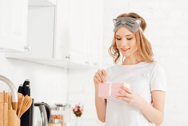 красивая девушка в пижаме и маске, открывающая подарочную коробку на кухне
 - Фото, изображение
