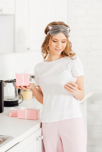 beautiful smiling girl in pajamas and sleeping mask holding gift box and using smartphone in kitchen - Photo, image