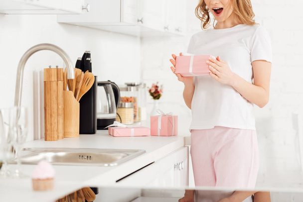 partial view of excited girl in pajamas holding gift box in kitchen - Фото, изображение