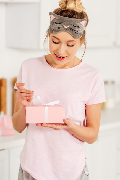 selective focus of beautiful smiling girl in pajamas and sleeping mask opening pink gift box at home - Φωτογραφία, εικόνα
