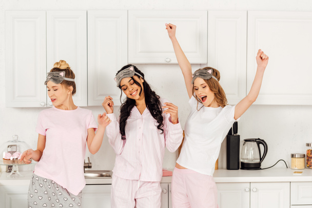 meninas multiculturais bonitas em máscaras de dormir dançando e se divertindo na cozinha durante a festa de pijama
 - Foto, Imagem