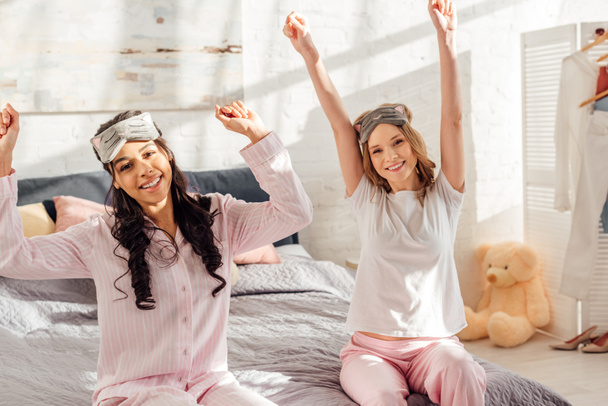 beautiful smiling multicultural girls looking at camera and stretching in morning during pajama party - Fotografie, Obrázek