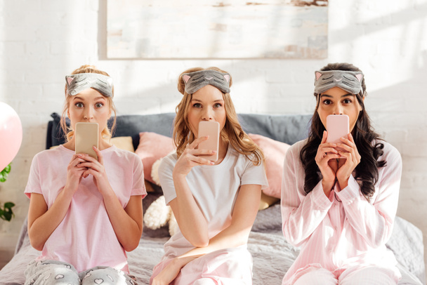 beautiful multicultural girls in sleeping masks sitting on bed and covering mouths with smartphones during pajama party - Photo, Image