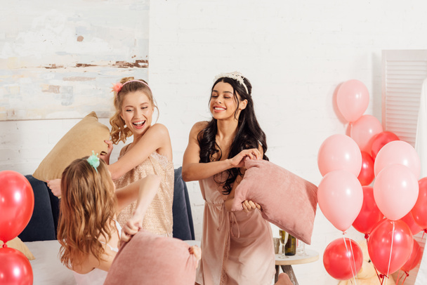 meninas multiétnicas bonitas em headbands se divertindo e lutando com travesseiros durante a festa de pijama no quarto
 - Foto, Imagem