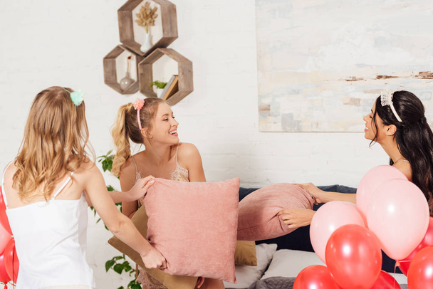 красивые мультиэтнические девушки в повязках веселятся и дерутся с подушками во время пижамной вечеринки в спальне
 - Фото, изображение