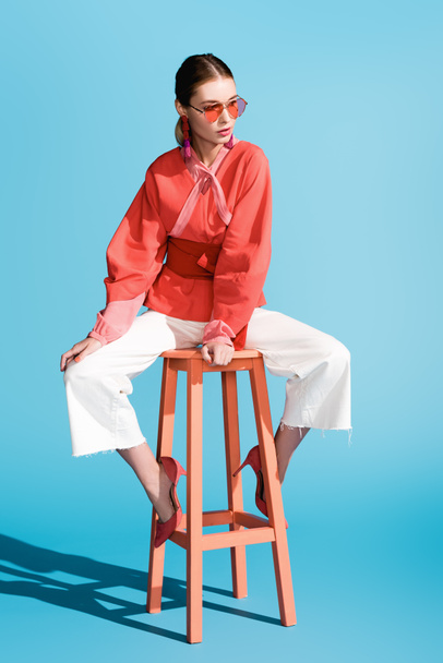 fashionable woman in living coral clothing and sunglasses posing on stool on blue - Photo, Image