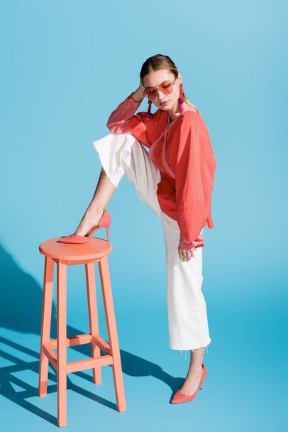 stylish model in living coral clothing and sunglasses posing on stool on blue - Photo, Image