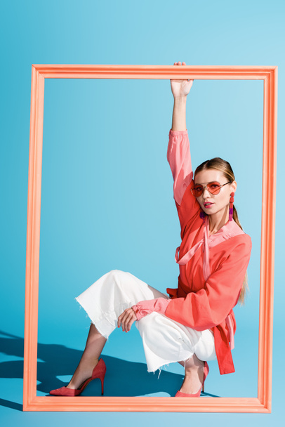 beautiful young woman in living coral clothing and sunglasses posing with big frame on blue - Fotó, kép