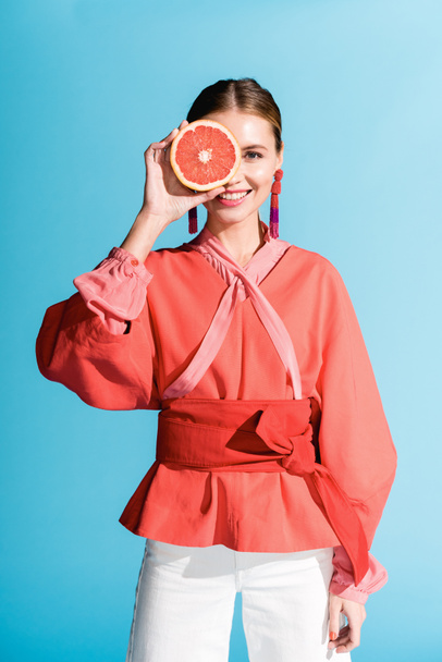 attractive smiling woman in living coral clothing posing with grapefruit isolated on blue  - Фото, изображение