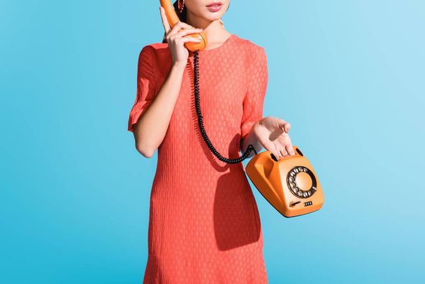 vista cortada da mulher em vestido de coral vivo posando com telefone rotativo isolado em azul
 - Foto, Imagem