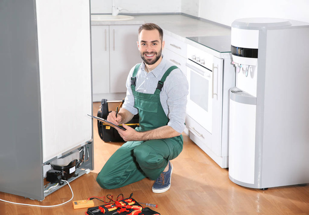 Male technician with clipboard and tools near broken refrigerator in kitchen - Foto, afbeelding