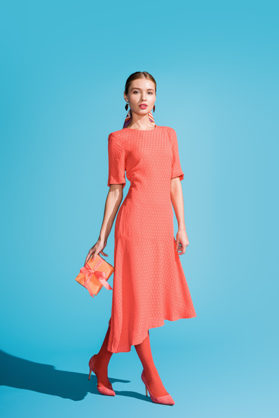 stylish woman in living coral dress holding gift box on blue - Fotografie, Obrázek