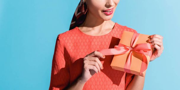 cropped view of fashionable woman in living coral dress holding gift box isolated on blue - Фото, зображення
