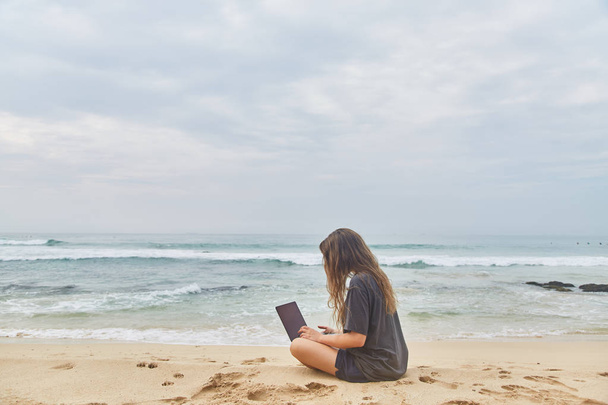 若い女の子がビーチでコンピューターで作業します。ビーチで働くフリーランサー. - 写真・画像