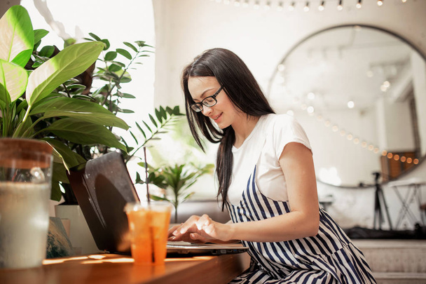 A pretty slim dark-haired girl with glasses,wearing casual style, types something on her laptop in a cozy coffee shop, - Foto, Imagen