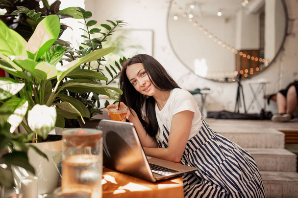 A beautiful young slim girl with dark hair and glasses,dressed in casual style, sits at the table with a laptop  in a new coffee shop. - Zdjęcie, obraz