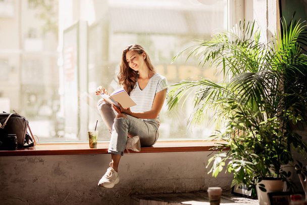 A pretty slim young girl with long hair,wearing casual outfit,sit on the windowsill  and reads a book in a cozy cafe. - Fotoğraf, Görsel