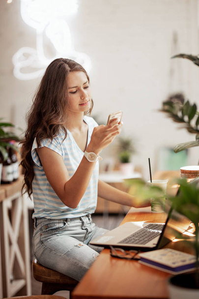 A young slim girl with long hair,wearing casual style, sits at the table with a laptop and looks at her phone in a cozy coffee shop. - Foto, imagen