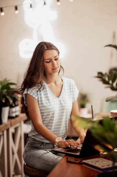 A young slim girl with long hair,wearing casual style, sits at the table with a laptop in a modern coffee shop. - Photo, image