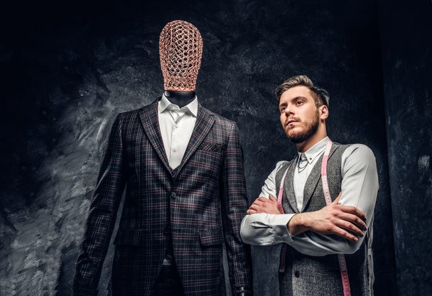 A confident young fashion designer posing next to a mannequin in exclusive custom made mens suit in a dark tailor studio  - Photo, Image