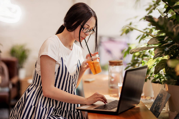 A beautiful young slim girl with dark hair and glasses,dressed in casual style, sits at the table with a laptop  in a new coffee shop. - 写真・画像
