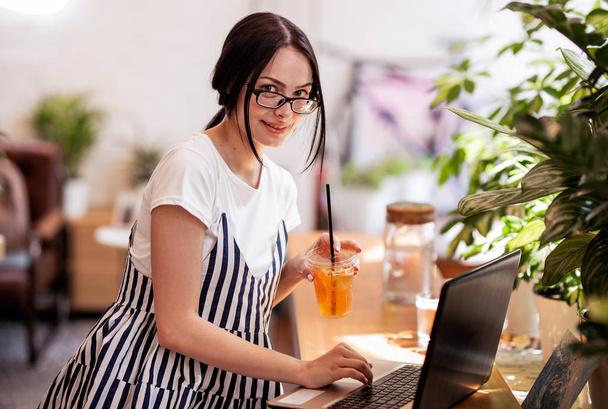 A beautiful young slim girl with dark hair and glasses,dressed in casual style, sits at the table with a laptop and looks at the camera in a cozy coffee shop. - Foto, Bild