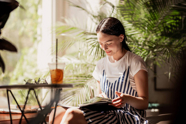 A young slim friendly girl with dark hair,dressed in casual outfit,sits at the table and reads a book in a cozy coffee shop. - Photo, image