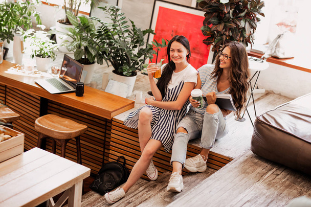 Two young smiling girls with long dark hair,wearing casual outfit,sit next to each other and drink coffee in a cozy coffee shop. . . - Foto, Imagem