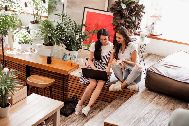 Two young smiling girls with long dark hair,wearing casual outfit,sit next to each other and drink coffee in a cozy coffee shop. - Foto, Imagem