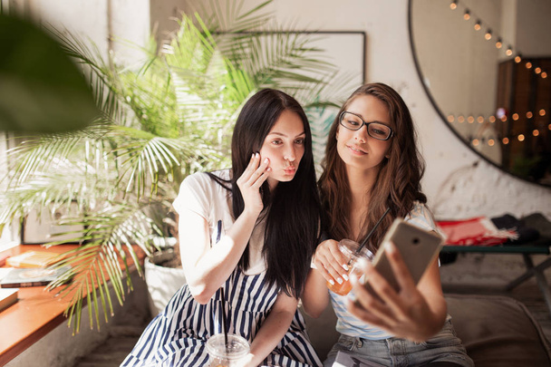 Two youthful smiling beautiful slim girls with long dark hair,wearing casual clothes, sit next to each other and take a selfie in a modern coffee shop. - Photo, image