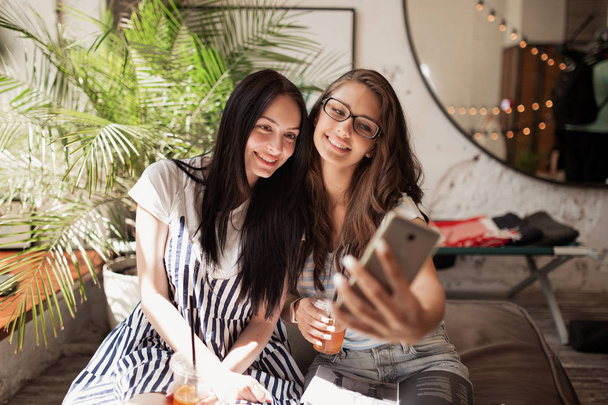 Two youthful smiling beautiful slim girls with long dark hair,wearing casual clothes, sit next to each other and take a selfie in a modern coffee shop. - Foto, immagini