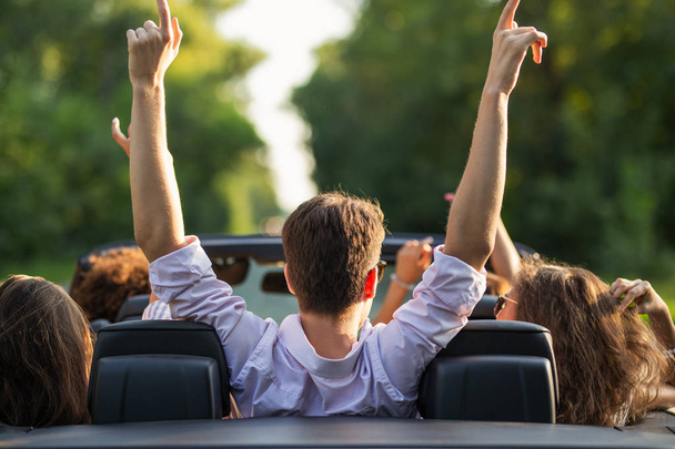Young man is sitting in a black cabriolet  between young women and holding their hands up on a sunny day. - Фото, изображение