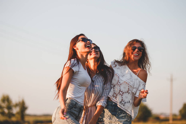Three gorgeous young women in sunglasses dressed in the beautiful clothes are laughing outdoor on a sunny day. - Foto, Imagem