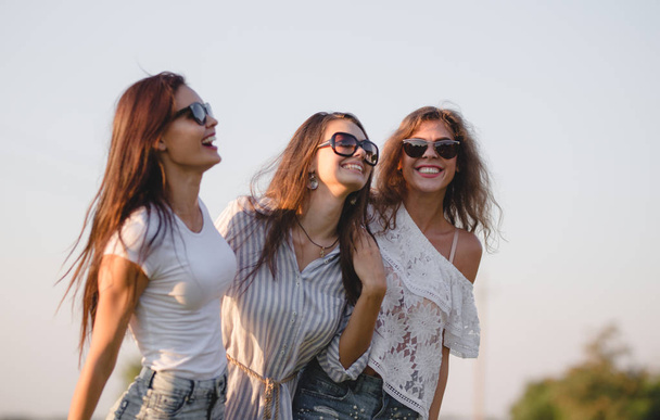Three gorgeous young women in sunglasses dressed in the beautiful clothes are laughing outdoor on a sunny day. - Foto, Bild
