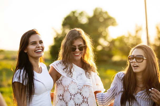 Three beautiful dark-haired young women in sunglasses dressed in the beautiful clothes are smiling and walking outdoor on a sunny day. - 写真・画像