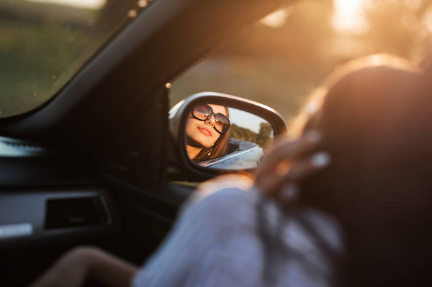 Reflection of a beautiful young dark-haired girl in sunglasses in a side mirror of a car. - Valokuva, kuva