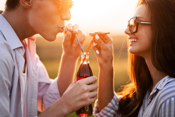 dunkelhaarige Mädchen und ein junger Mann trinken an einem sonnigen Tag aus einer Flasche ein Getränk durch Strohhalme im Freien. - Foto, Bild