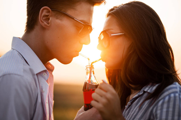 Beautiful dark-haired girl and a young man  in sunglasses are drinking from one bottle a drink through straws outdoor on a sunny day. - Photo, image