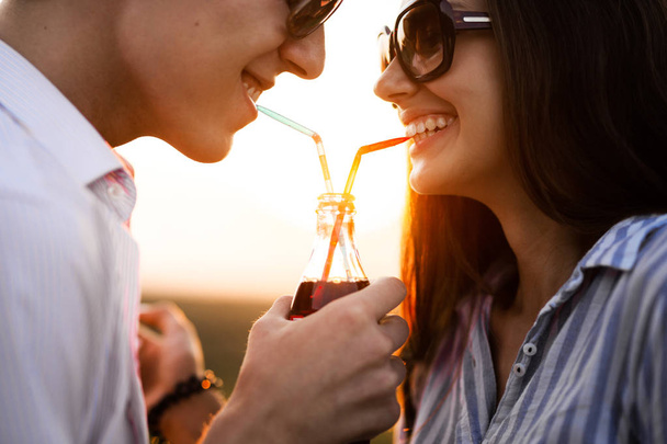 Beautiful dark-haired girl and a young man  in sunglasses are drinking from one bottle a drink through straws outdoor on a sunny day. - Φωτογραφία, εικόνα